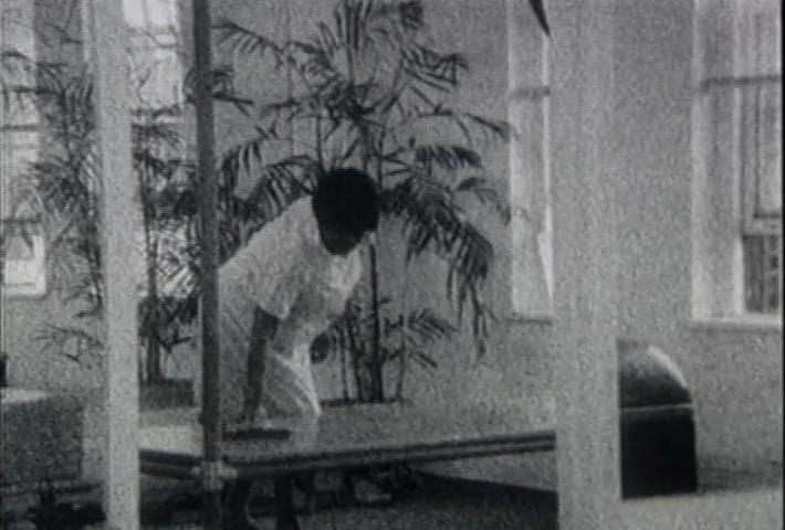 A black and white still from Fannie's film showing a black woman in a white cleaner's uniform wiping a coffee table. There are white walls and large indoor plants in the background.