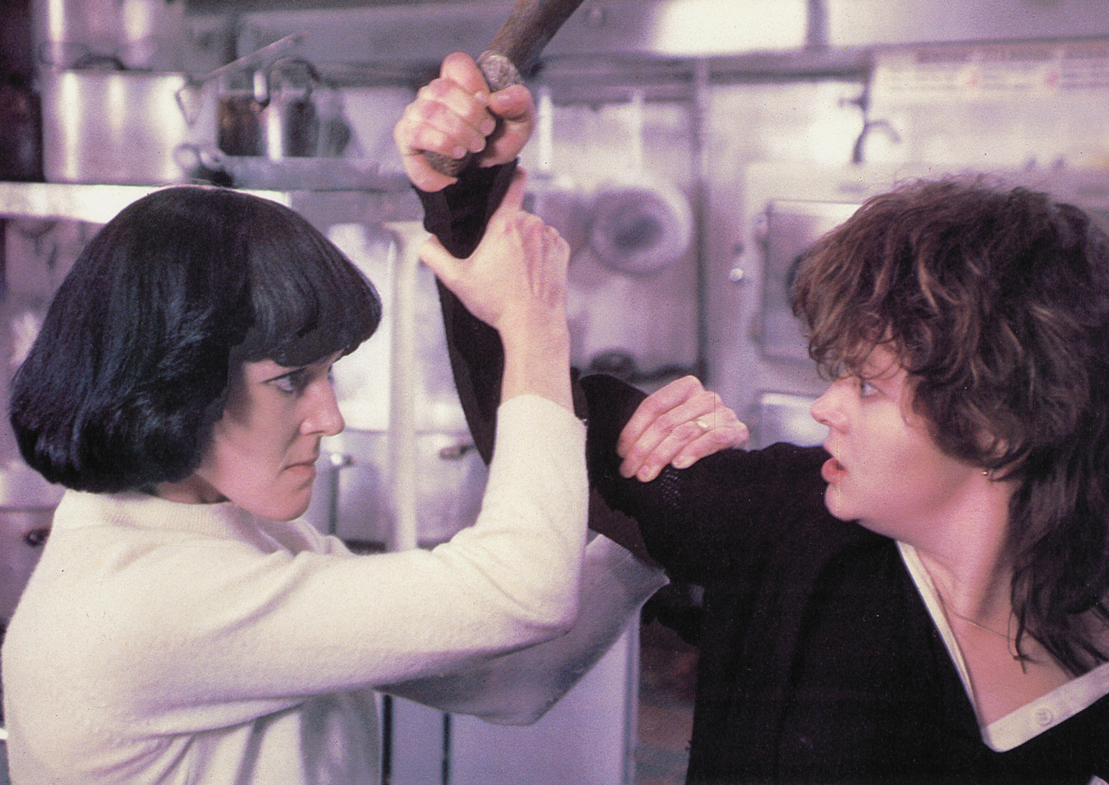 Two women stand together in a combative pose. The one on the left, wearing a white jumper, holds the right's arm. The one on the right, wearing a black jumper, raises a wooden club in their right arm.
