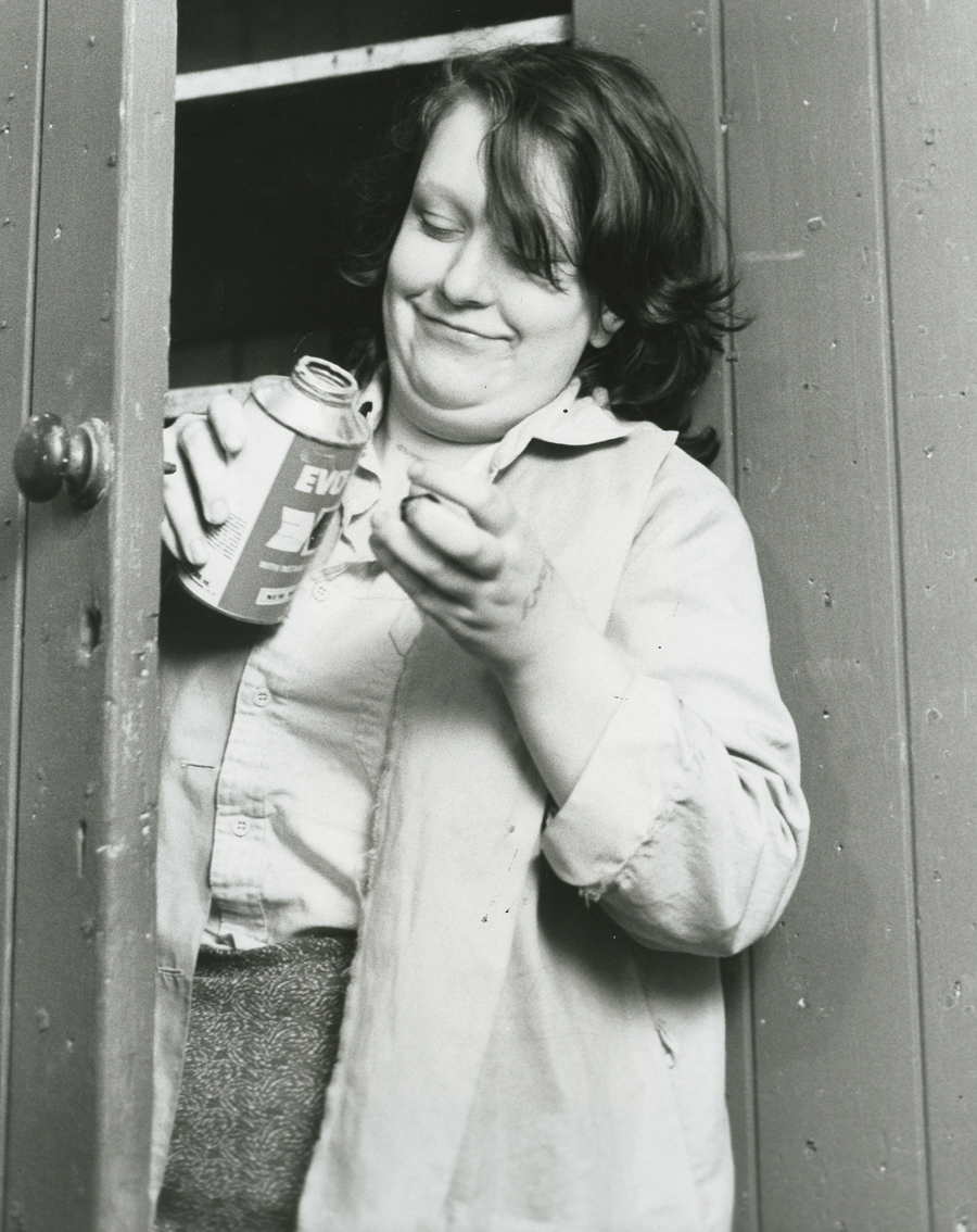 A black and white image of a woman in a white coat smiling as she opens a bottle of some liquid (perhaps paint).