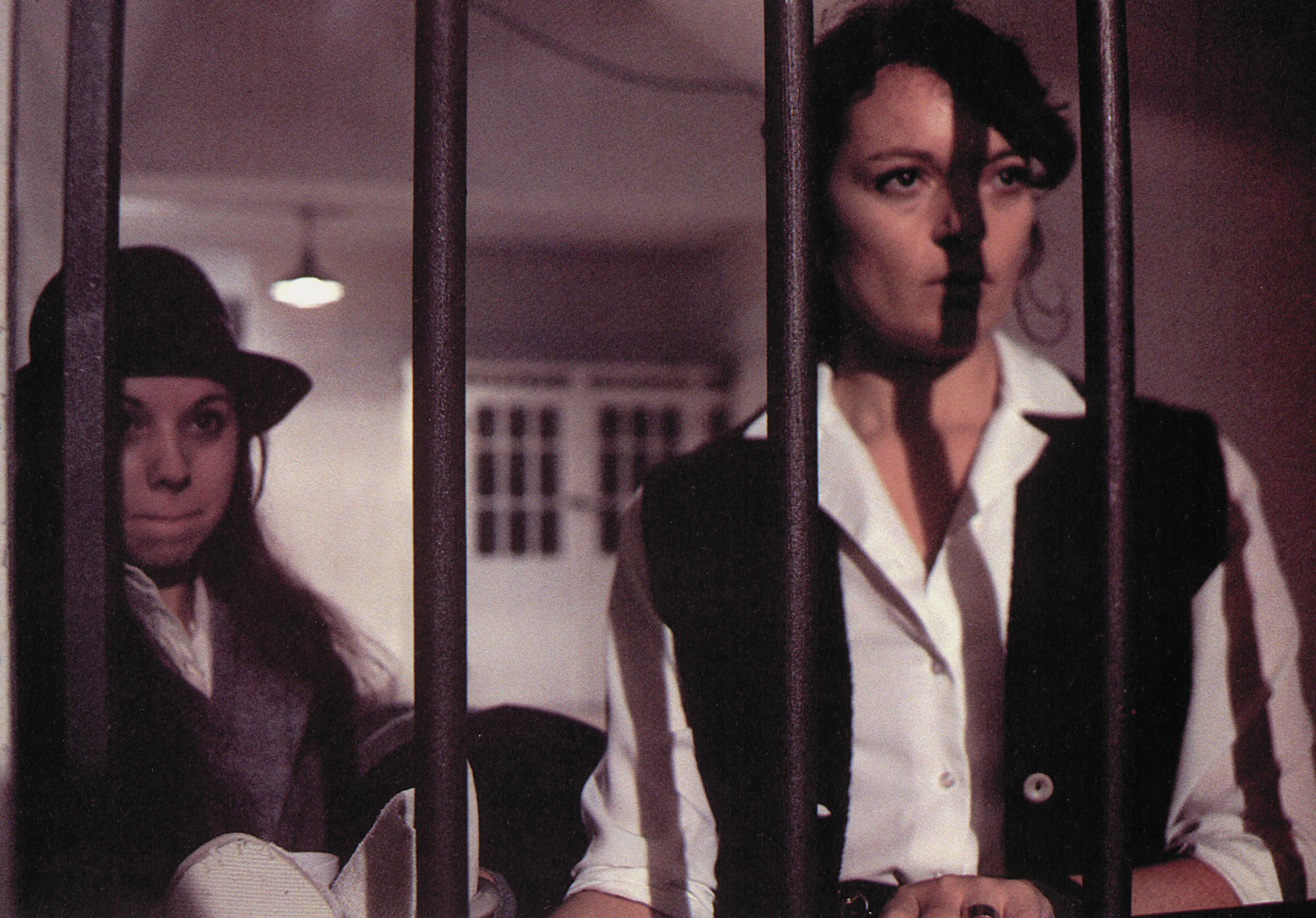 Two women stand behind bars in a prison cell. The one on the right wears a white shirt and black sleeveless jacket. A shadow from one of the cell bars is cast upon her face.