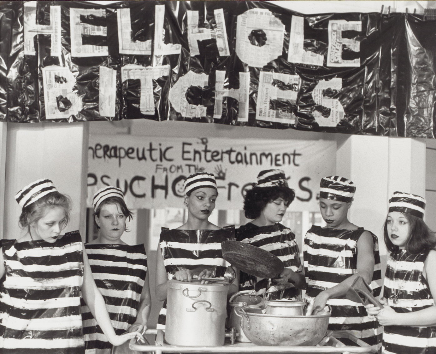 A black and white image of six women wearing plastic sheets (striped with white paint to look like prison garments) and standing in front of some kitchen utensils. A sign above their heads reads 'Hellhole Bitches'