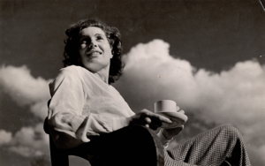 A black and white photo of a woman sitting back in a wooden chair while holding a cup and saucer. The image is shot from below and behind her we can see clouds passing in the sky.