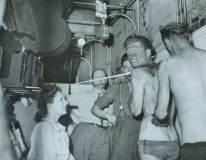 A black and white behind the scenes photo of a young woman on the set of a film. On the right are a line of coal miners, topless and covered in dirt. On the left the young woman smiles up at them from her seat.