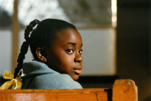 A young girl sits on a wooden bench, her head turned to the right to look over her shoulder.