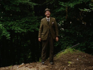 An elderly man wearing a hat and green jacket, stands on a earthy mound in a wood.