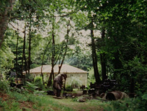 A man works on a machine in a wooded clearing. In the background is a house.