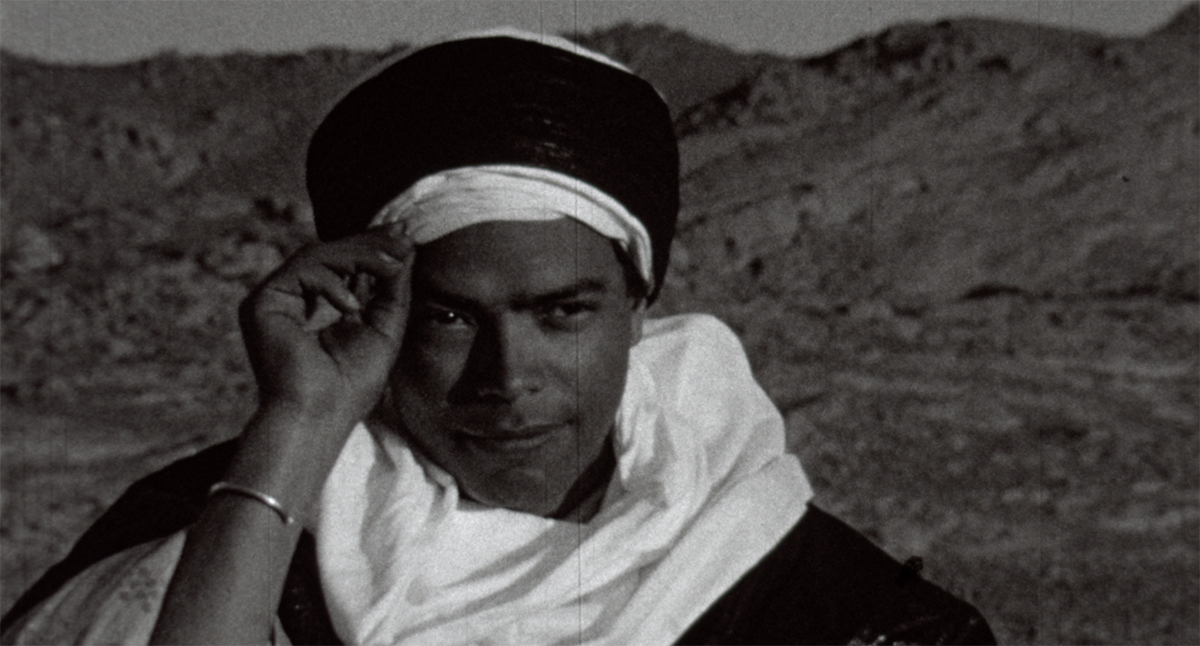 A black and white image showing a person standing in a desert with a white scarf and holding a black and white headscarf to their head with their right hand.