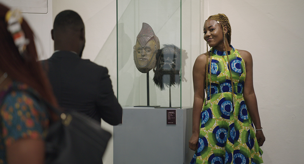 A person in a blue and green patterned dress poses for a photo next to an African mask in a glass cabinet.