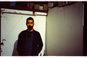 A person with a dark beard and short dark hair stands for a photo in front of a white exterior wall. They wear a blue shirt and navy jacket, and stand with their hands in the jacket pockets.